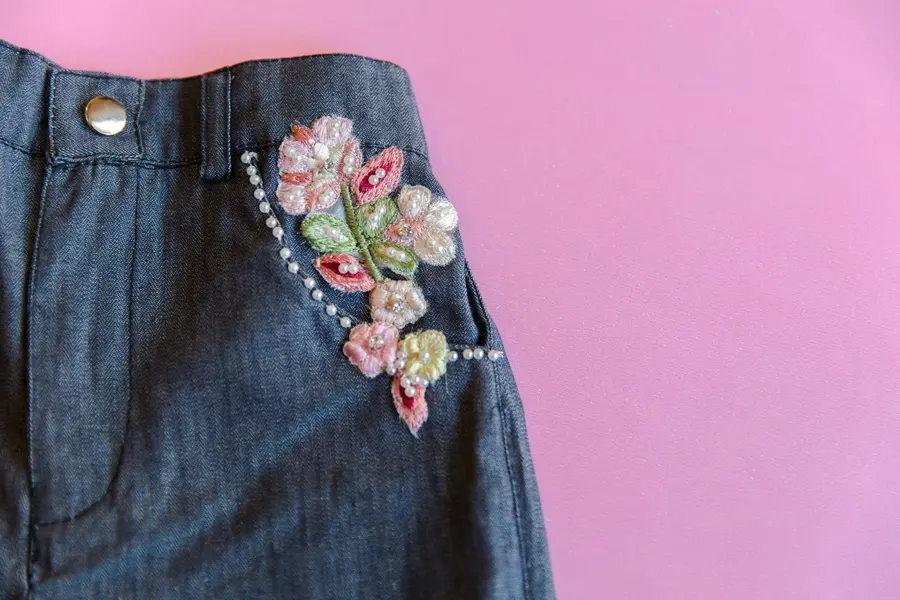 Pink and green floral embroidery with pearl beads on the front pocket of dark denim, on a pink background.