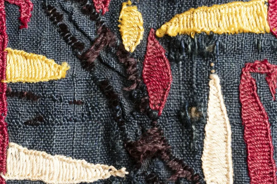 A close-up of thick yellow, red, cream and brown satin stitches on coarse dark blue woven fabric.
