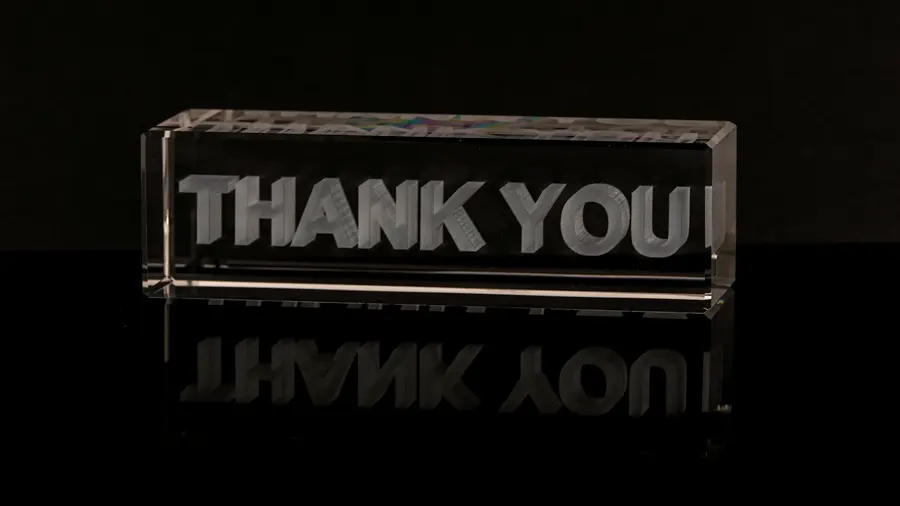 A clear crystal glass block with the words thank you engraved inside it, reflected on a black surface.