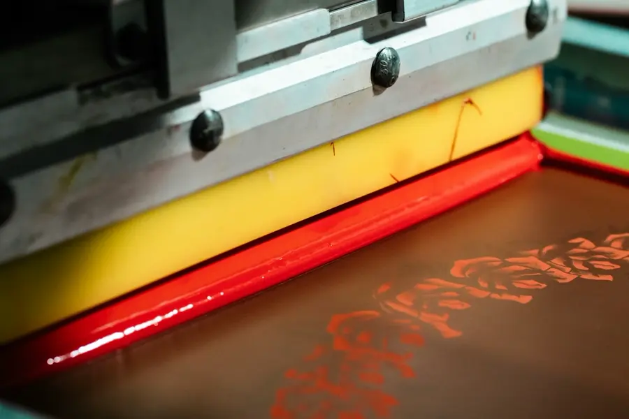 A close-up of a press squeegee pushing a bead of red ink across a screen with a floral stencil.