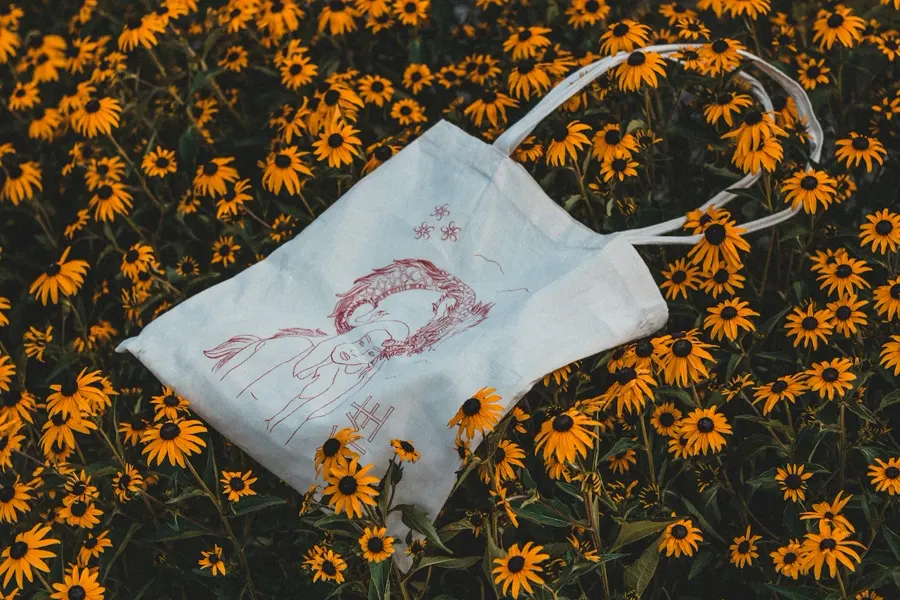 A natural cotton tote printed with a red line drawing of a figure and a dragon, lying on black-eyed Susan flowers.