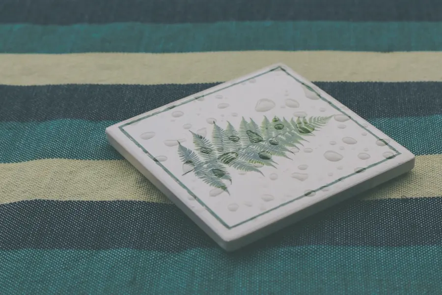 A square white ceramic coaster printed with a green fern frond and border, beaded with water drops, on striped fabric.