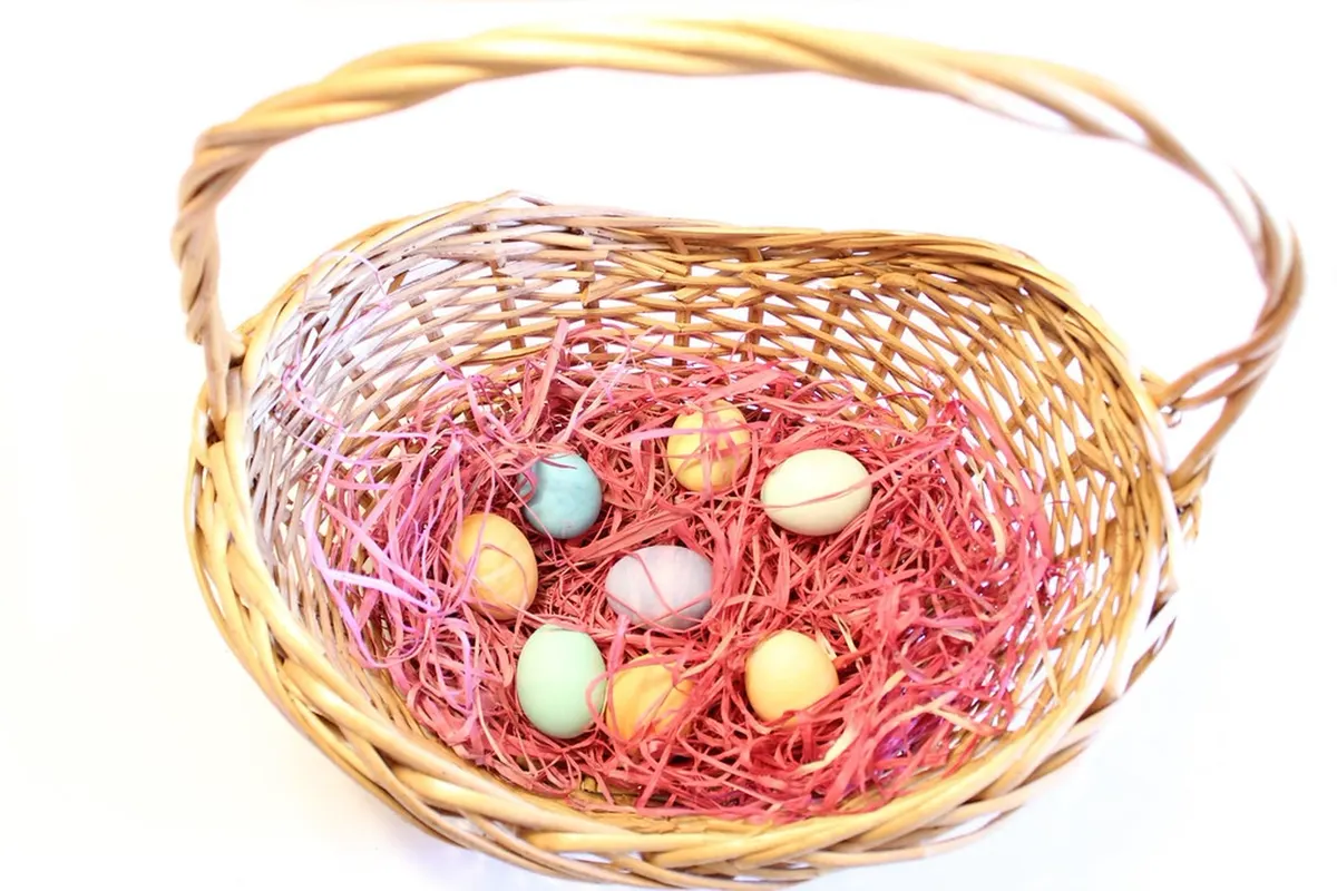 A wicker Easter basket with a tall handle, filled with pink paper grass and pastel eggs, on white.