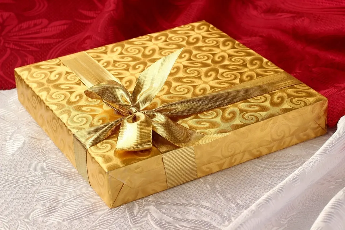 A flat gift box wrapped in embossed gold swirl paper and tied with a gold satin ribbon bow, on white lace with red fabric behind.