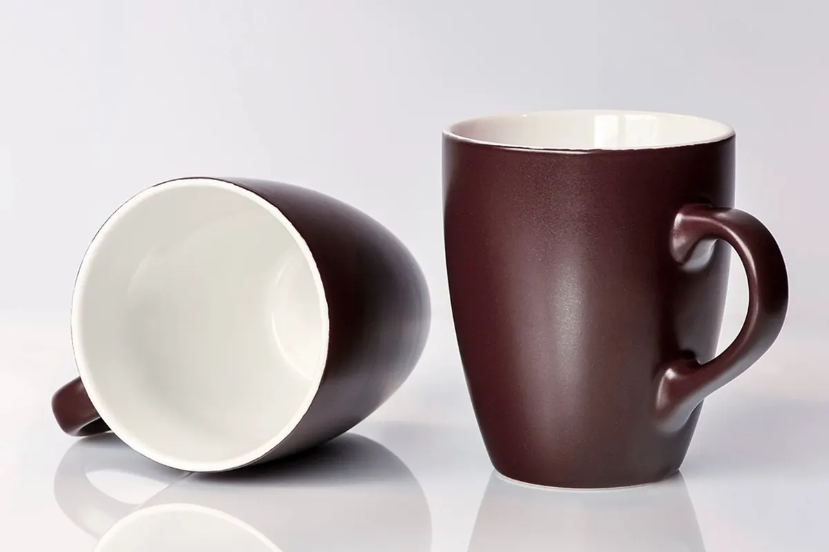 Two matte plum ceramic mugs with white interiors, one upright and one on its side, on a white reflective surface.