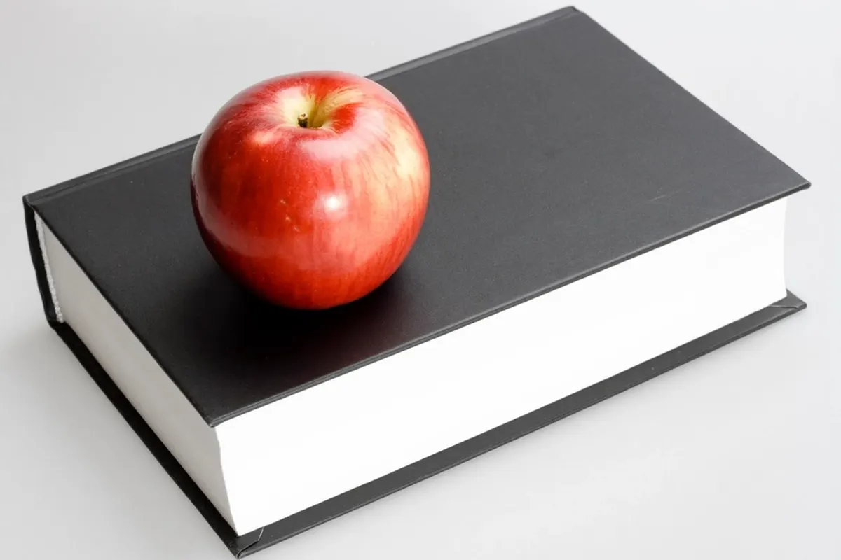 A shiny red apple resting on a closed black hardback book with white page edges, on a white surface.