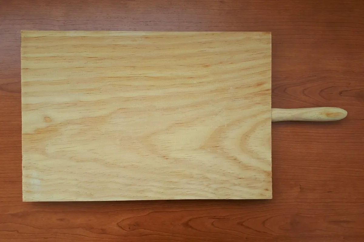 A plain light pine serving board with a round handle lying on a dark cherry wood table.