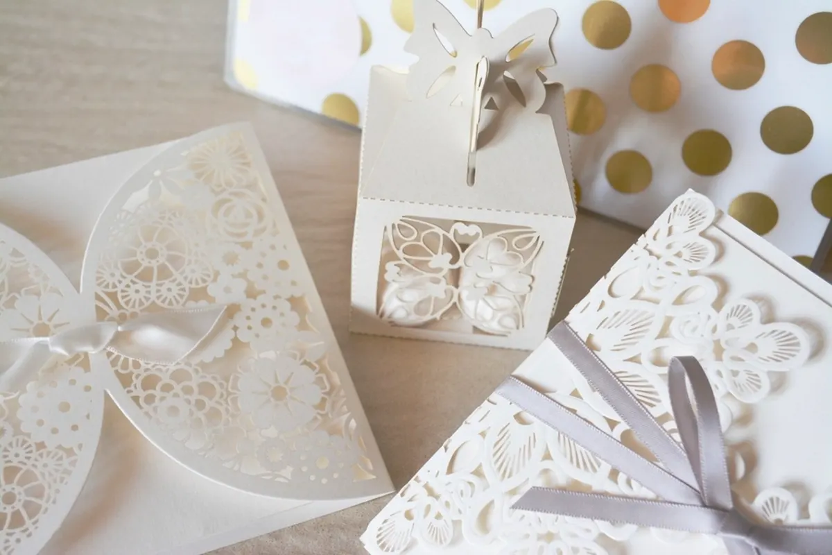 White laser-cut lace wedding favour boxes and invitations tied with grey satin ribbon, beside gold polka-dot paper.